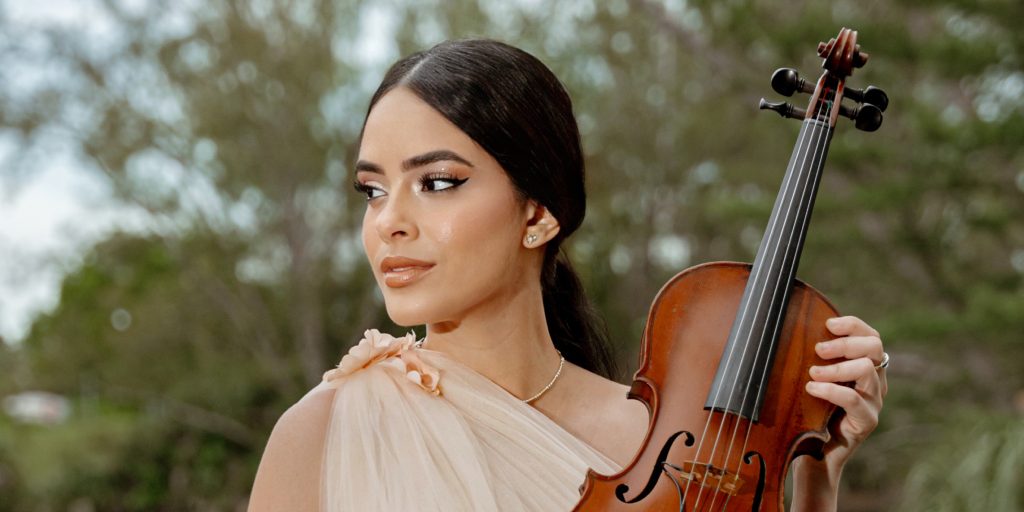 Maggi vi standing on a wooden deck in an elegant cream gown, holding a violin outdoors.