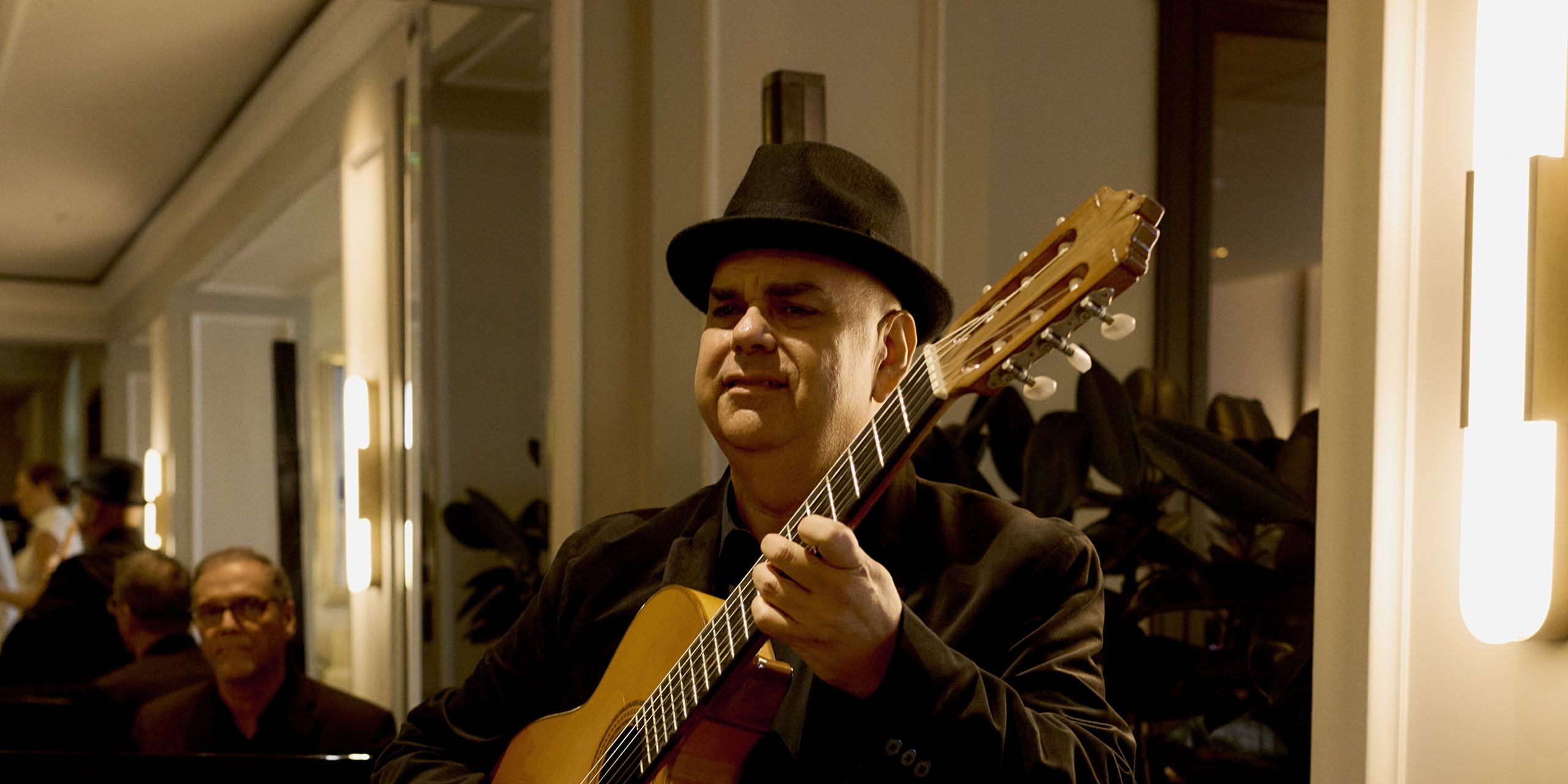 Man in a black hat playing guitar indoors with a pianist performing in the background.