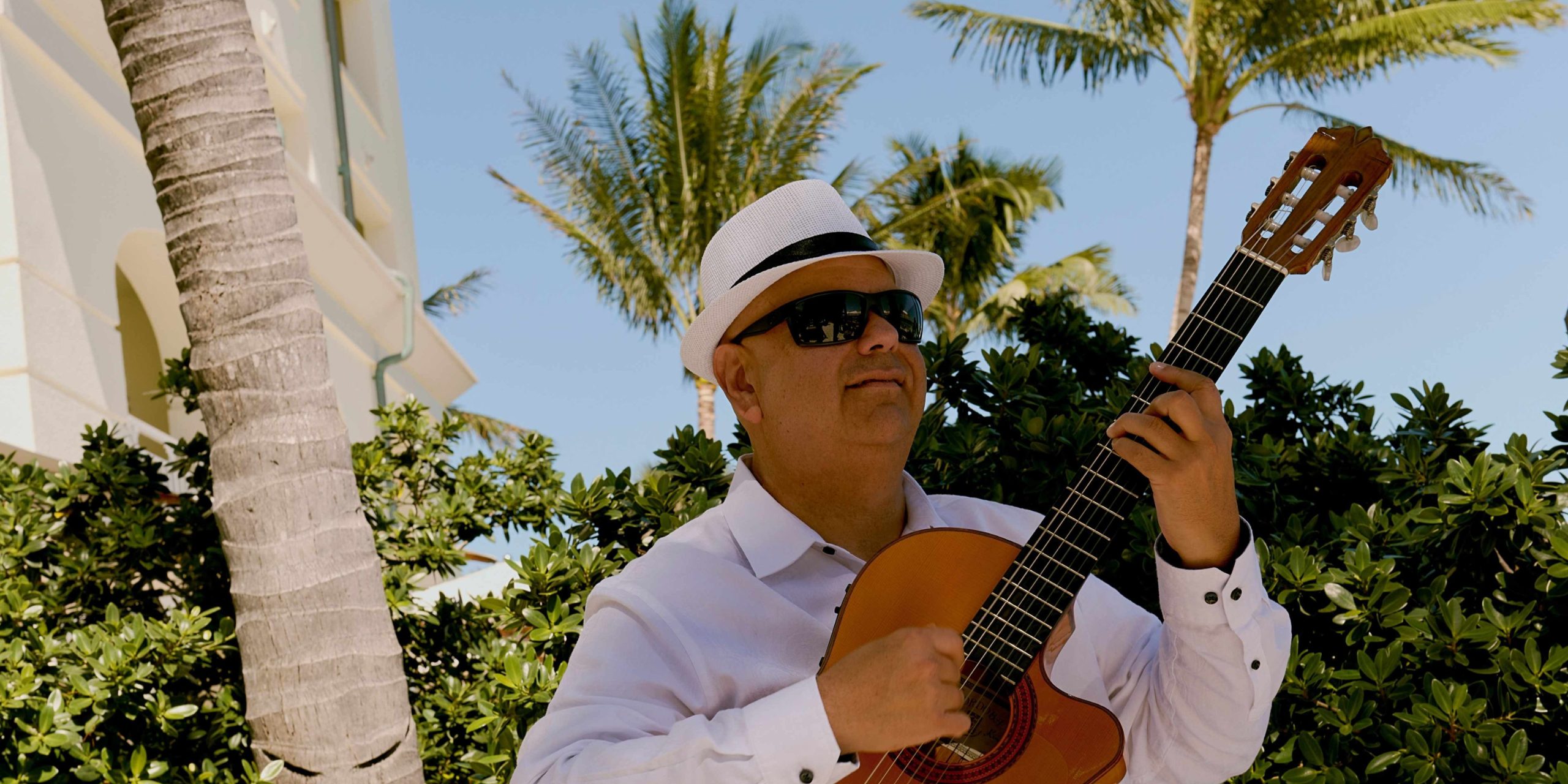 Person in a white hat and sunglasses playing an acoustic guitar outdoors with palm trees in the background.