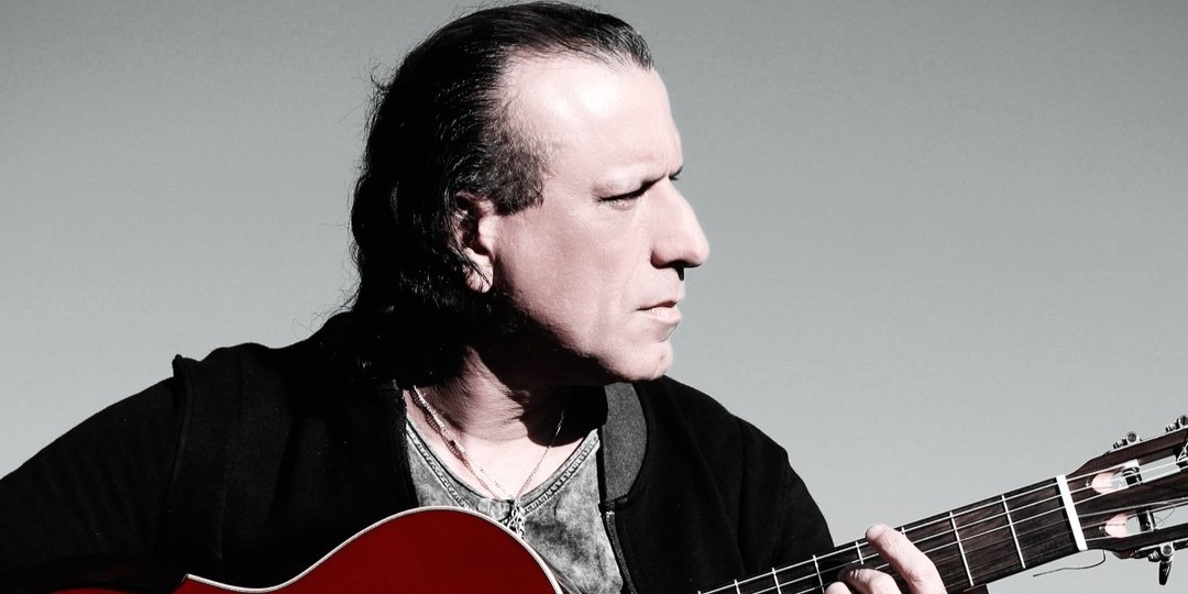 Person playing a red‑bodied classical guitar, looking to the side against a soft gray background.