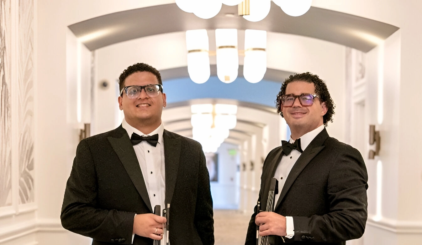 Two members of Cadenza The Group in tuxedos holding electric violins in an elegant hallway.