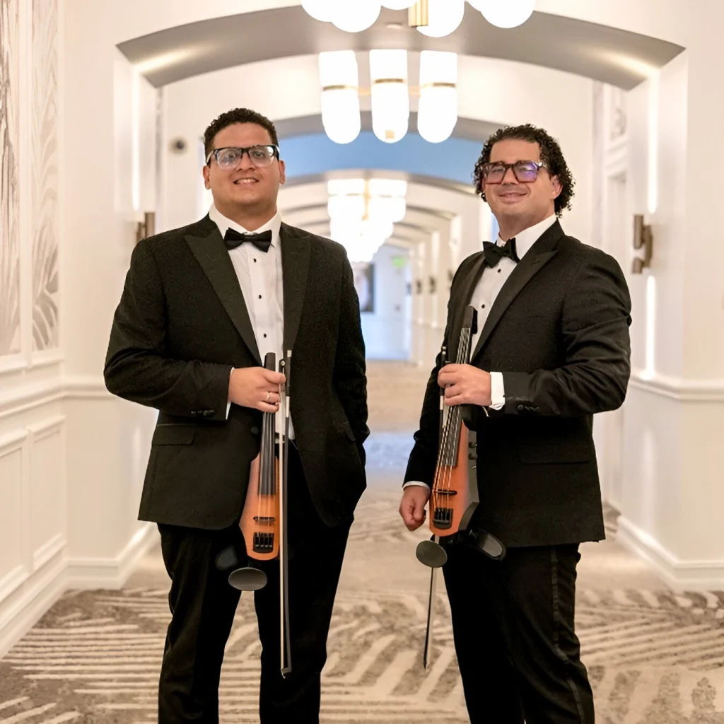 Two members of cadenza the group in tuxedos holding electric violins in an elegant hallway.