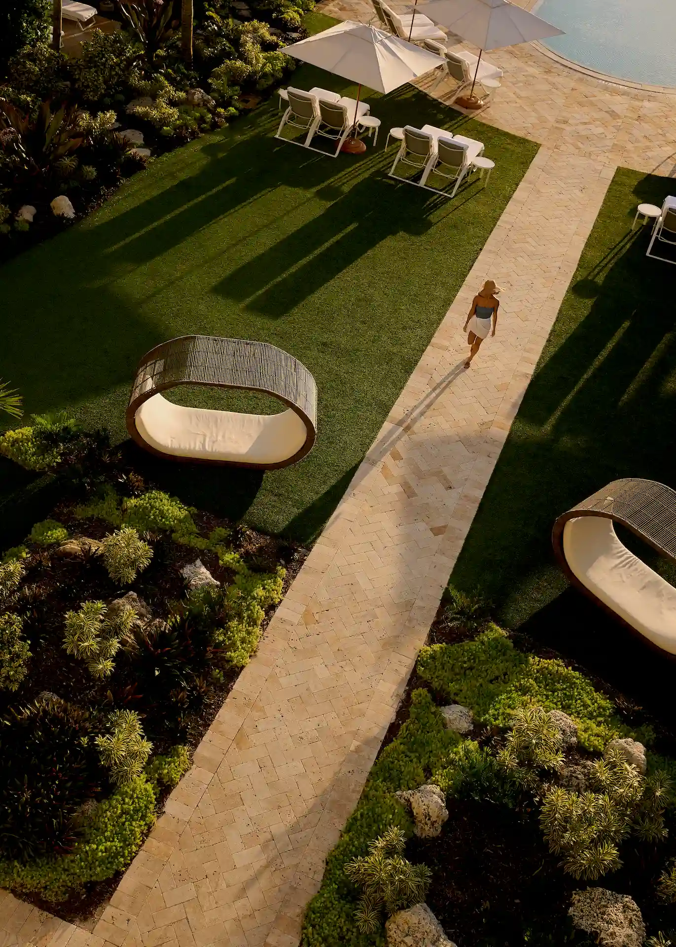 Stone walkway through lush gardens with modern loungers and poolside chairs under warm afternoon light.