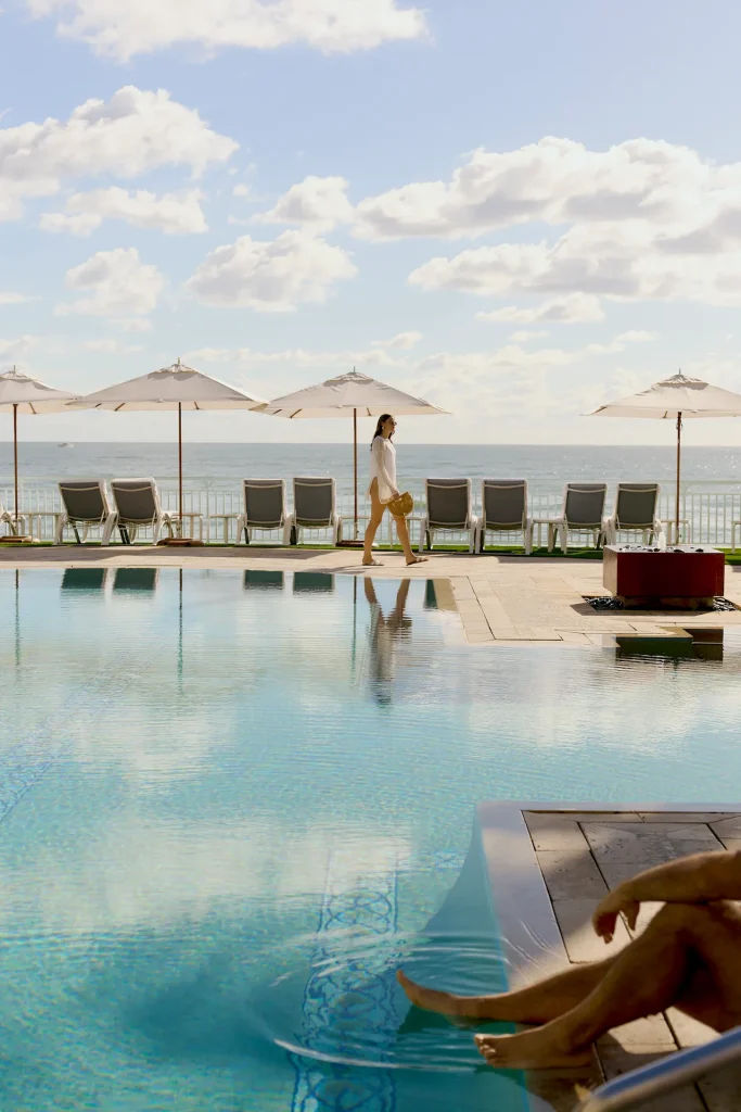 Woman walking by a calm oceanfront pool while another person relaxes with feet in the water.