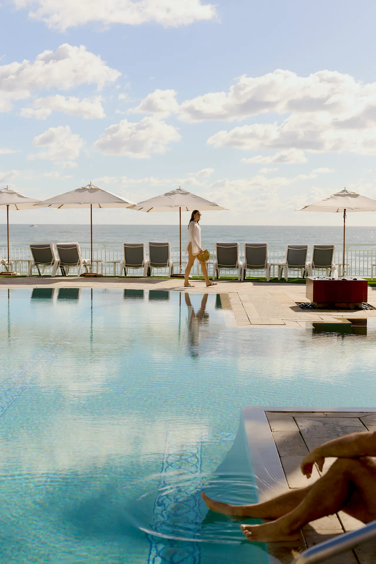 Woman walking by a calm oceanfront pool while another person relaxes with feet in the water.