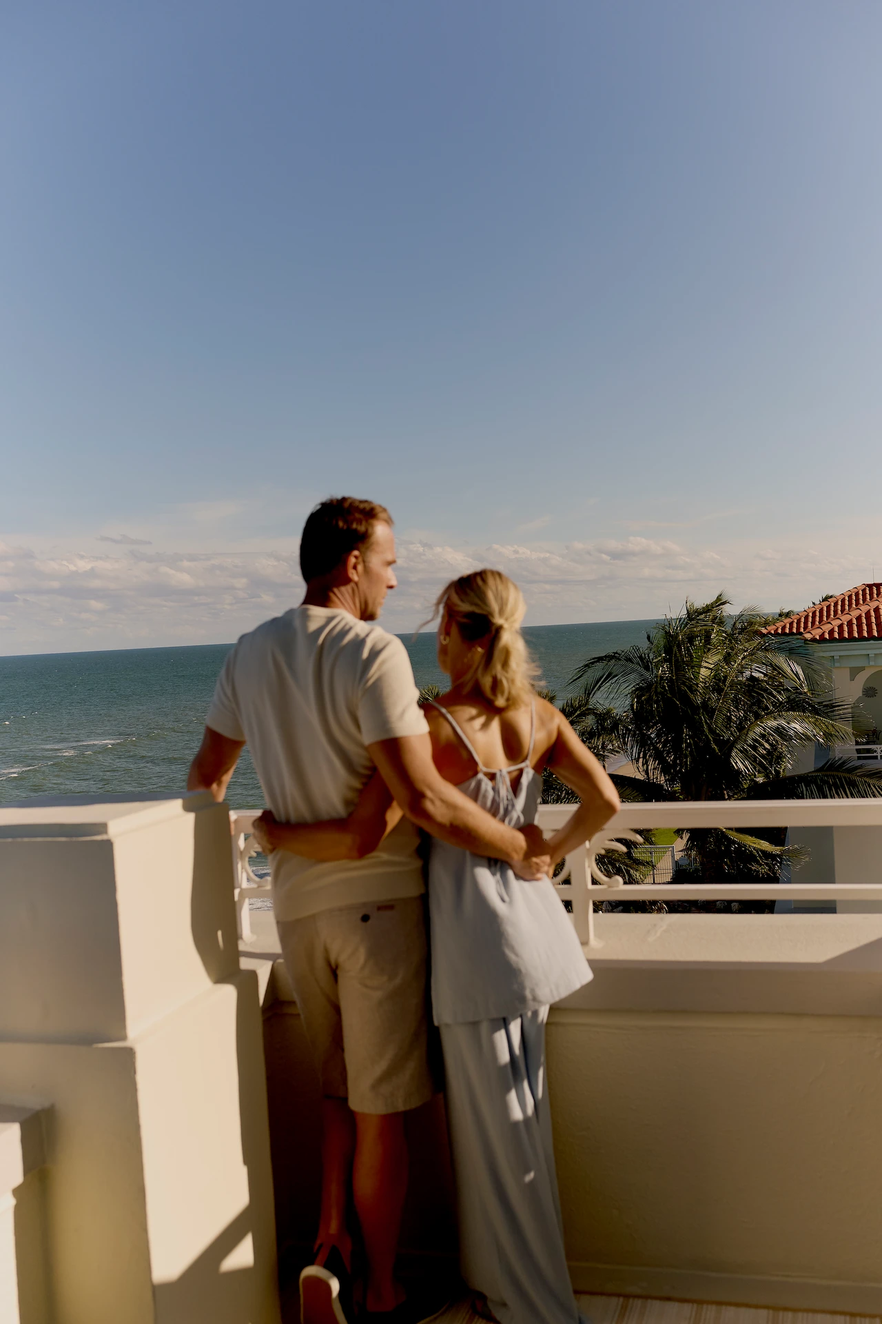 Couple embracing on a balcony overlooking the ocean with palm trees and clear blue sky.