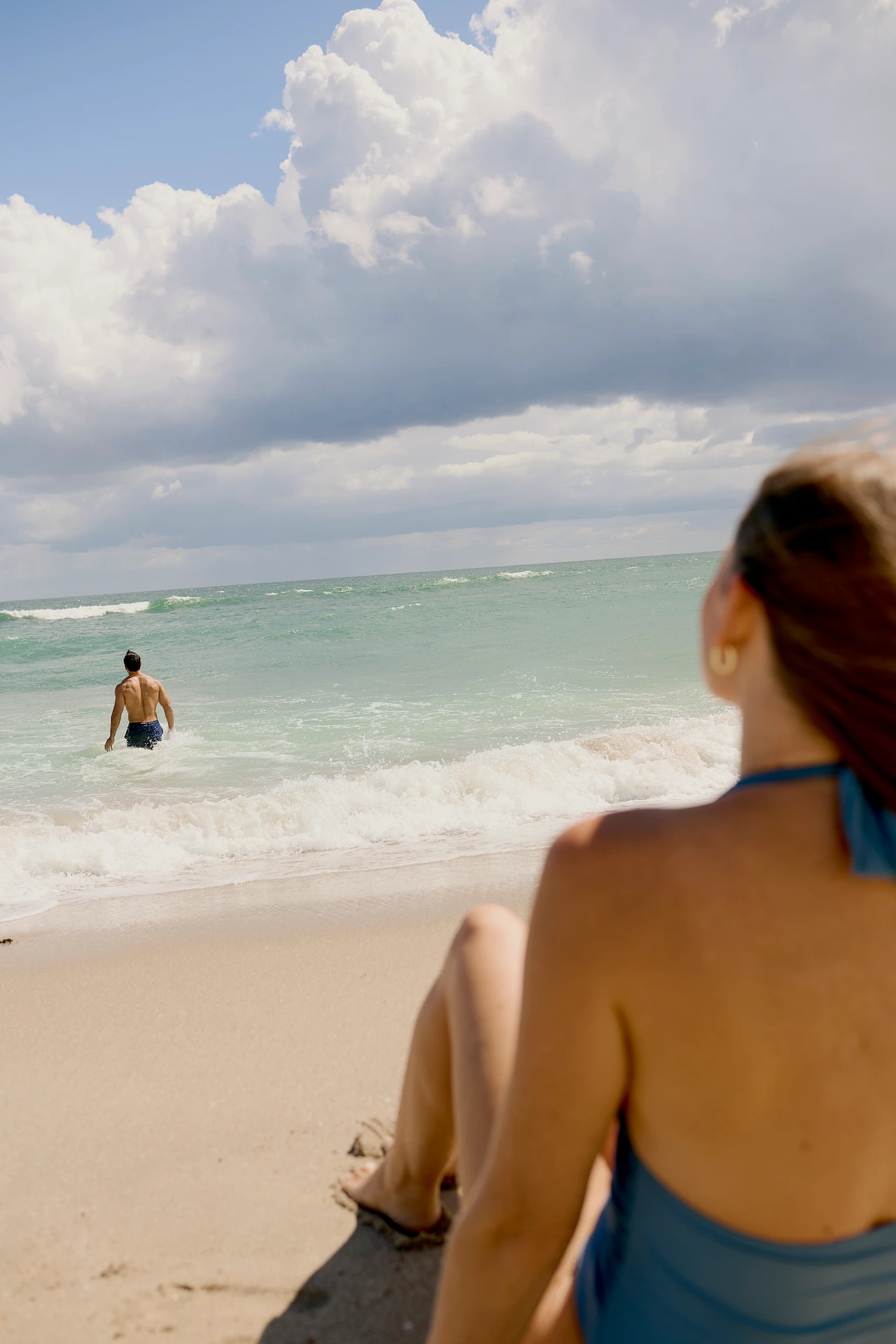 Person sitting on the sand watching someone walk into the ocean under a dramatic cloudy sky.