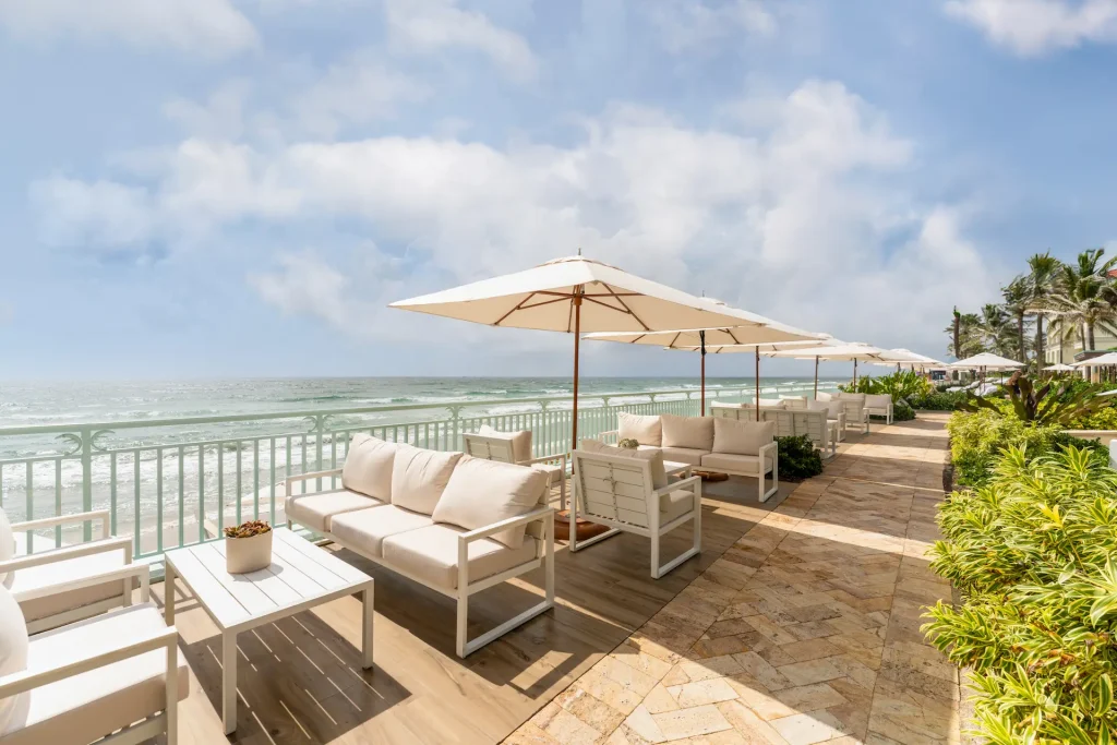Oceanfront patio at eau palm beach with white cushioned seating, large umbrellas, and tropical greenery overlooking the waves.