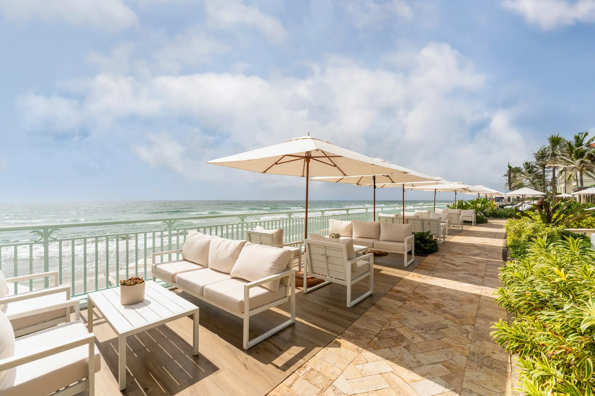 Oceanfront patio at eau palm beach with white cushioned seating, large umbrellas, and tropical greenery overlooking the waves.