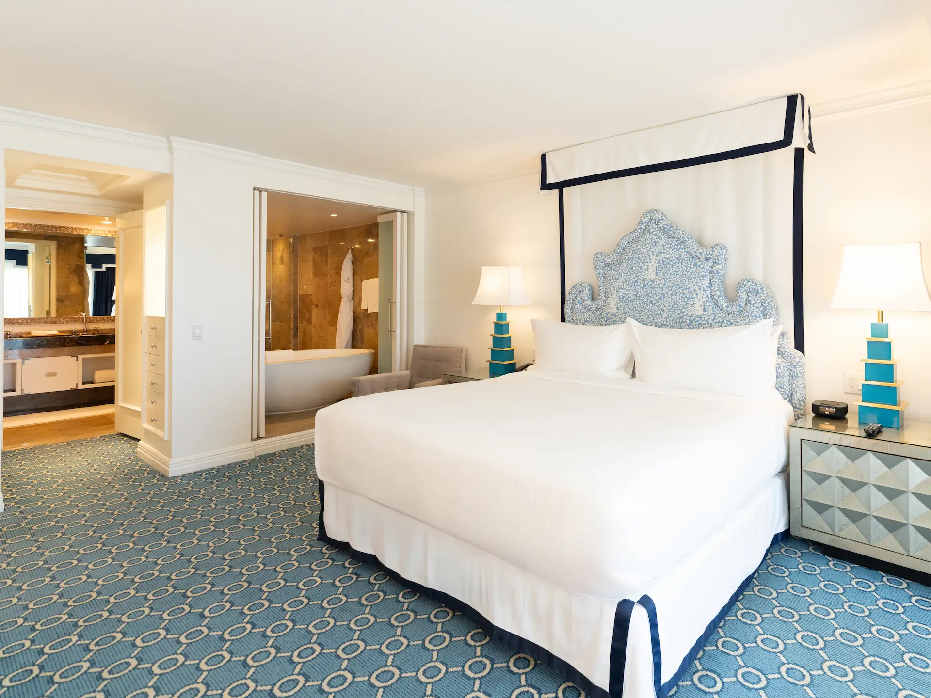 Suite bedroom with blue‑and‑white canopy headboard, white linens, patterned carpet, and view into bathroom.