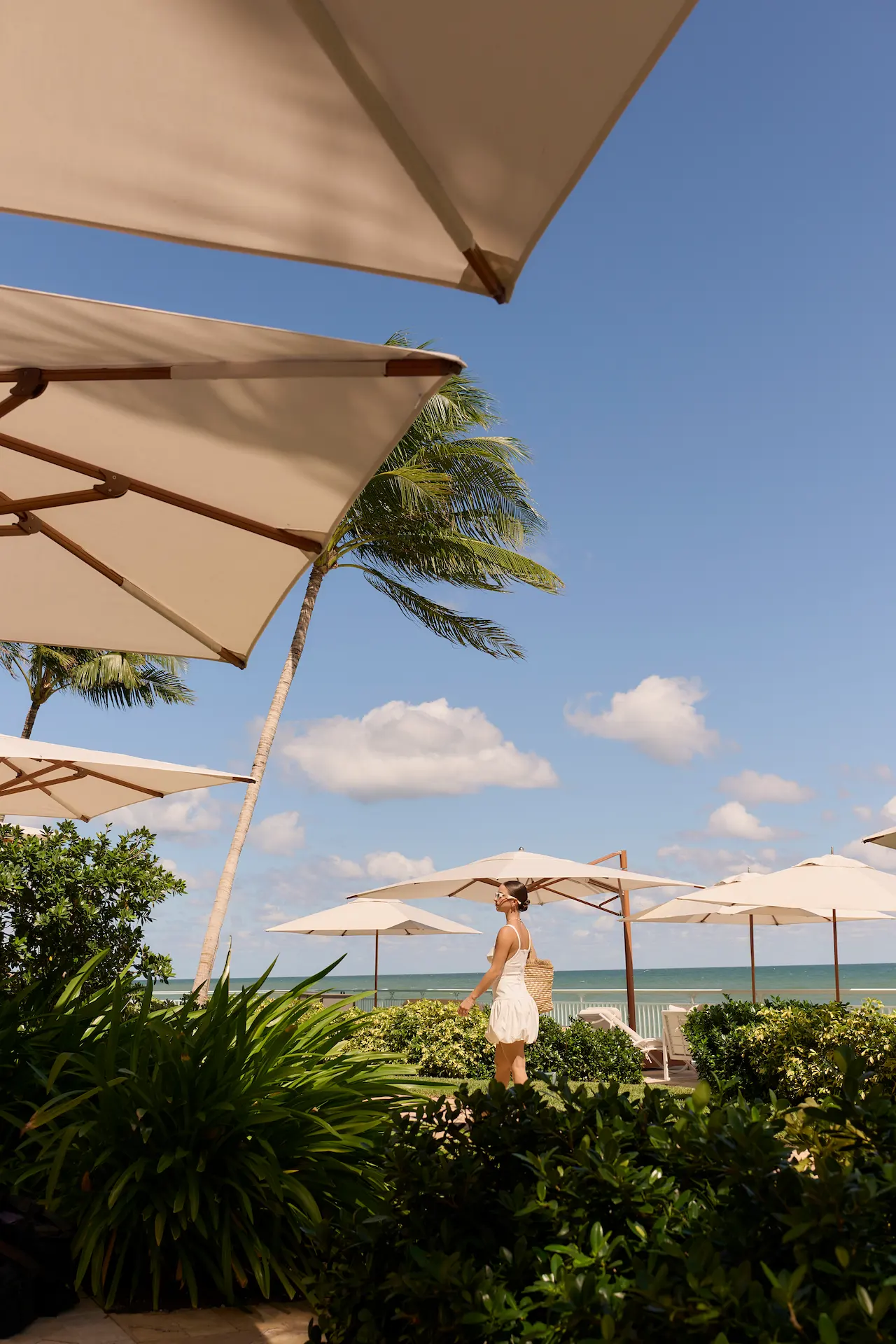 Person walking among palm trees and white umbrellas near the ocean.