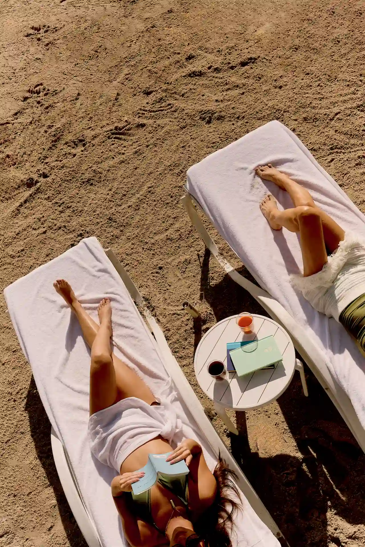 Two people relaxing on sunbeds with a small table holding drinks and a book.