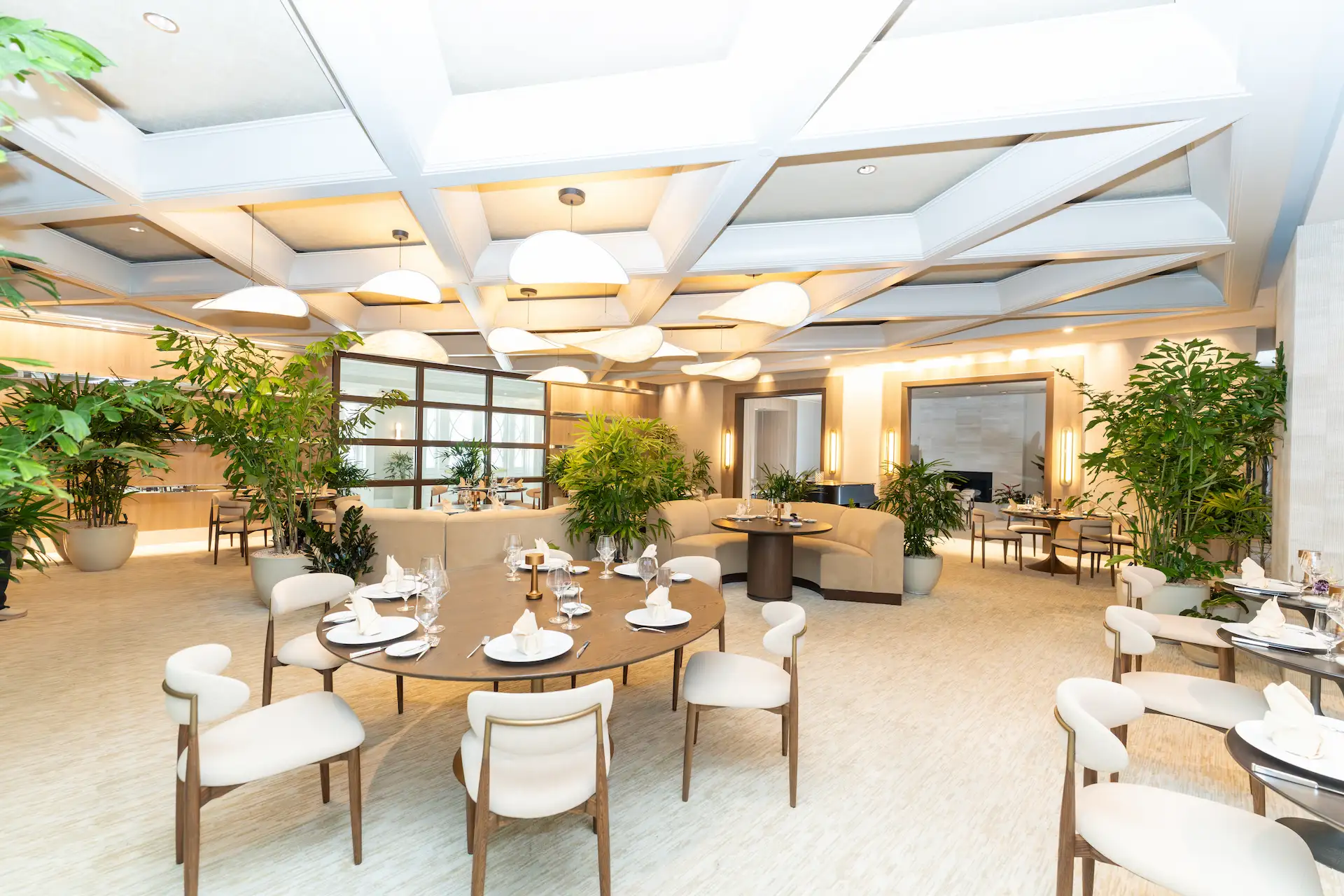 Upscale dining room with round tables, soft lighting, and large potted plants.