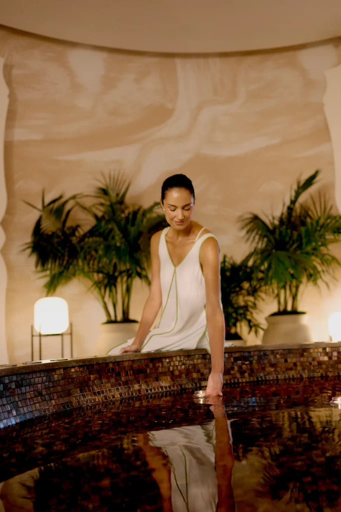 A woman sitting by an indoor water feature, gently touching the surface as soft lighting and greenery create a calm atmosphere.
