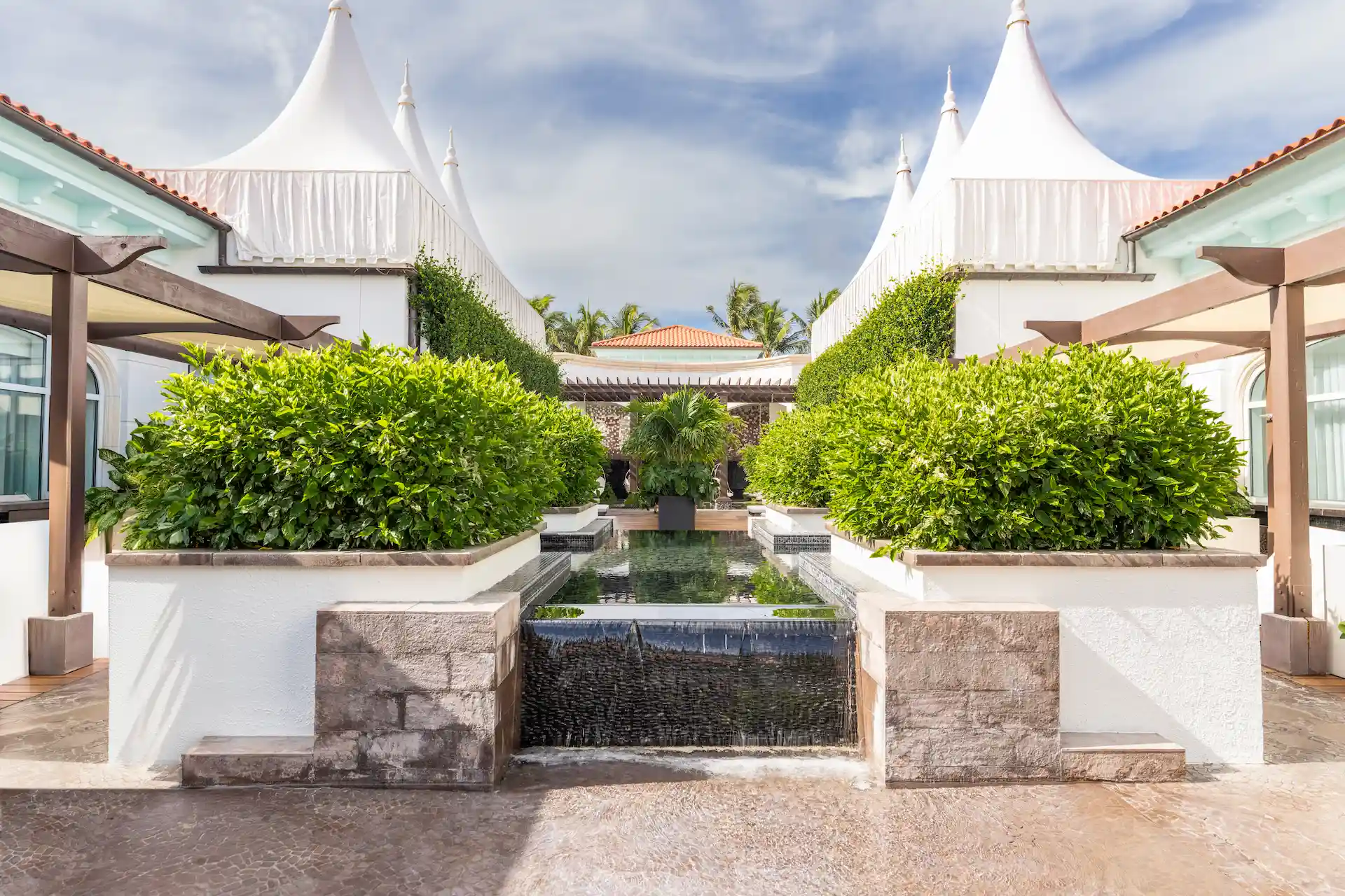 A landscaped courtyard with hedges, a central water feature, and white structures under a clear sky.