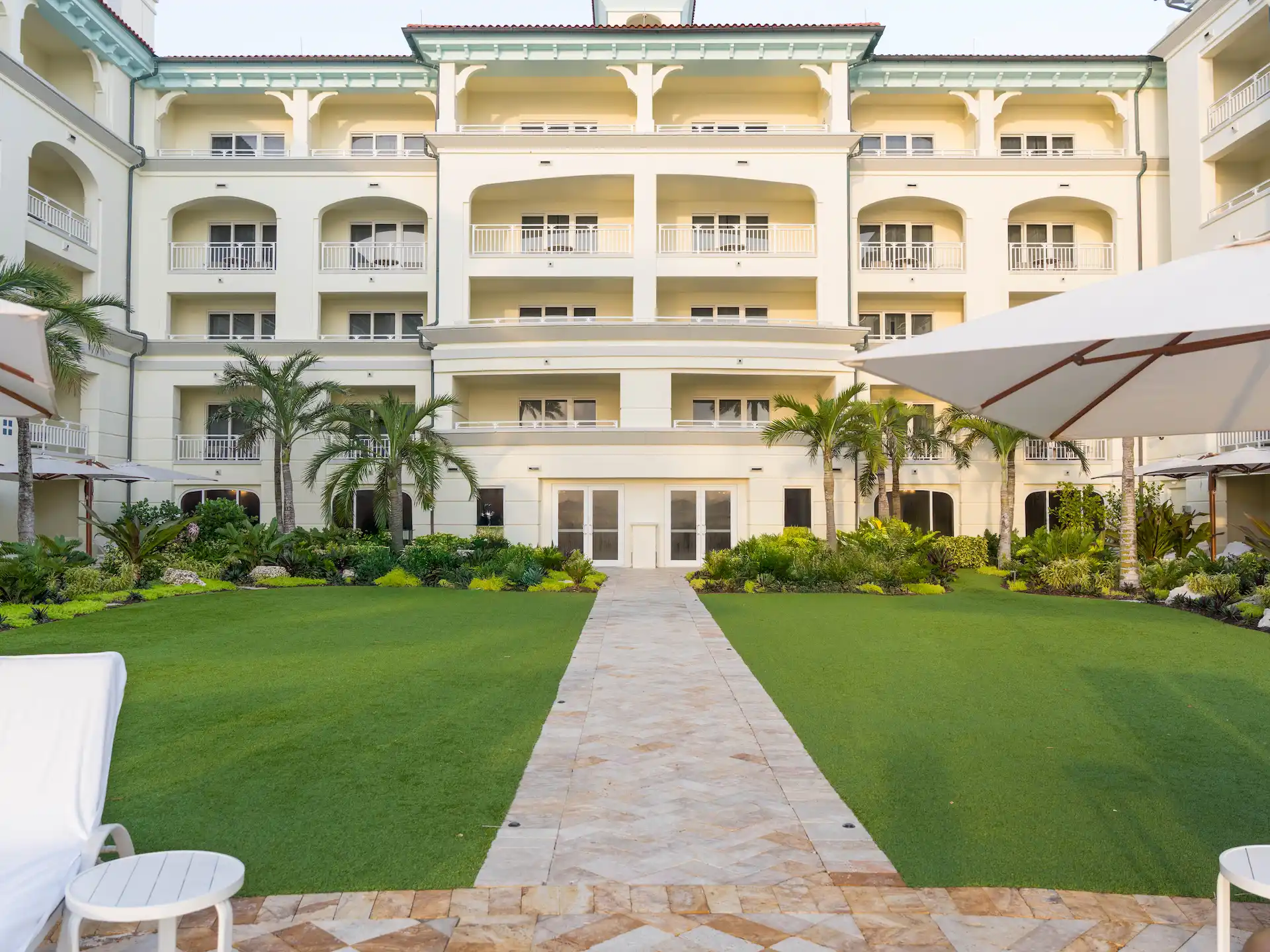 A resort façade with balconies overlooking a landscaped garden, palm trees, and outdoor seating areas.