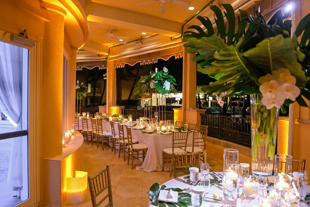 Banquet tables with tropical centerpieces, gold chairs, and warm lighting in an indoor‑outdoor setting.