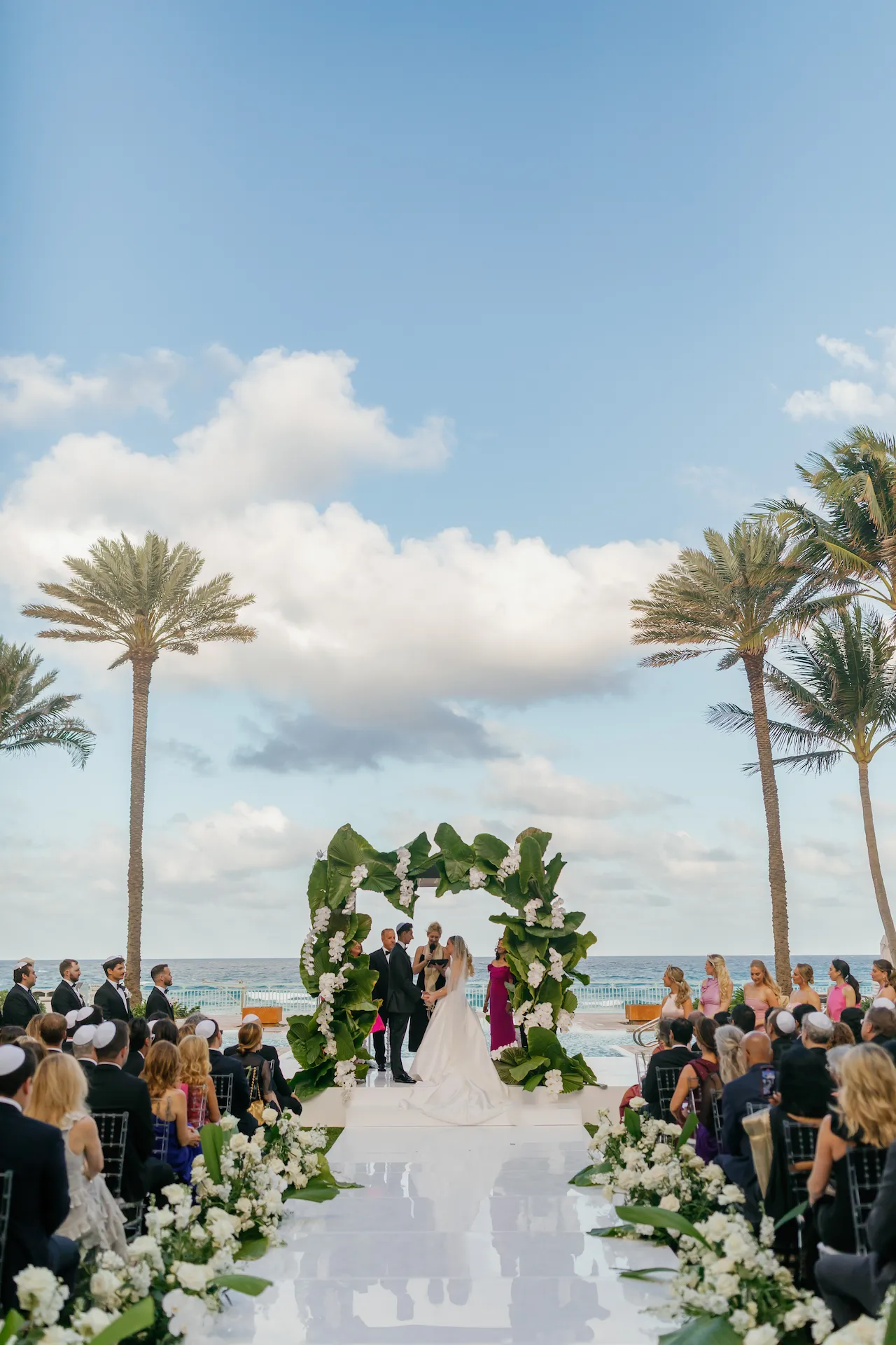 Beachside wedding ceremony with a floral arch, guests seated along the aisle, and ocean views.