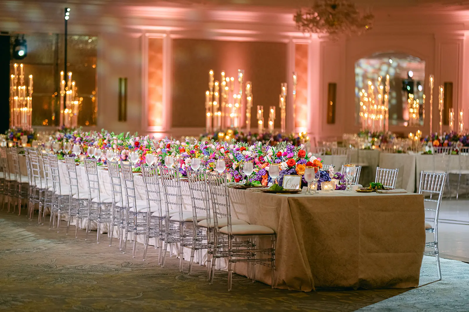 Banquet hall with long tables, colorful floral centerpieces, candles, and transparent chairs.