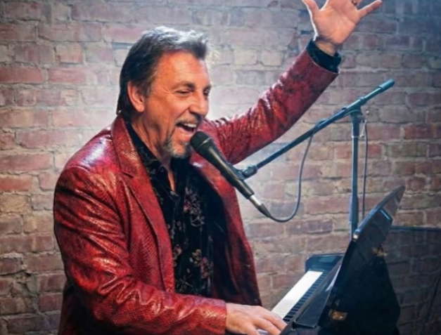 Man in a shiny red jacket performing with a microphone raised in front of a brick wall.