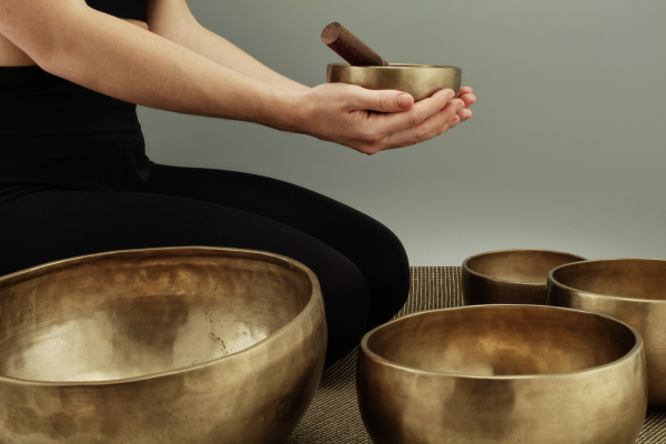 Person seated on the floor holding a brass singing bowl, with bowls arranged on a mat in front against a plain background