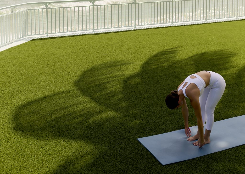 Person in a white athletic outfit doing a forward bend on a yoga mat outdoors, with ocean and palm‑leaf shadows behind them.
