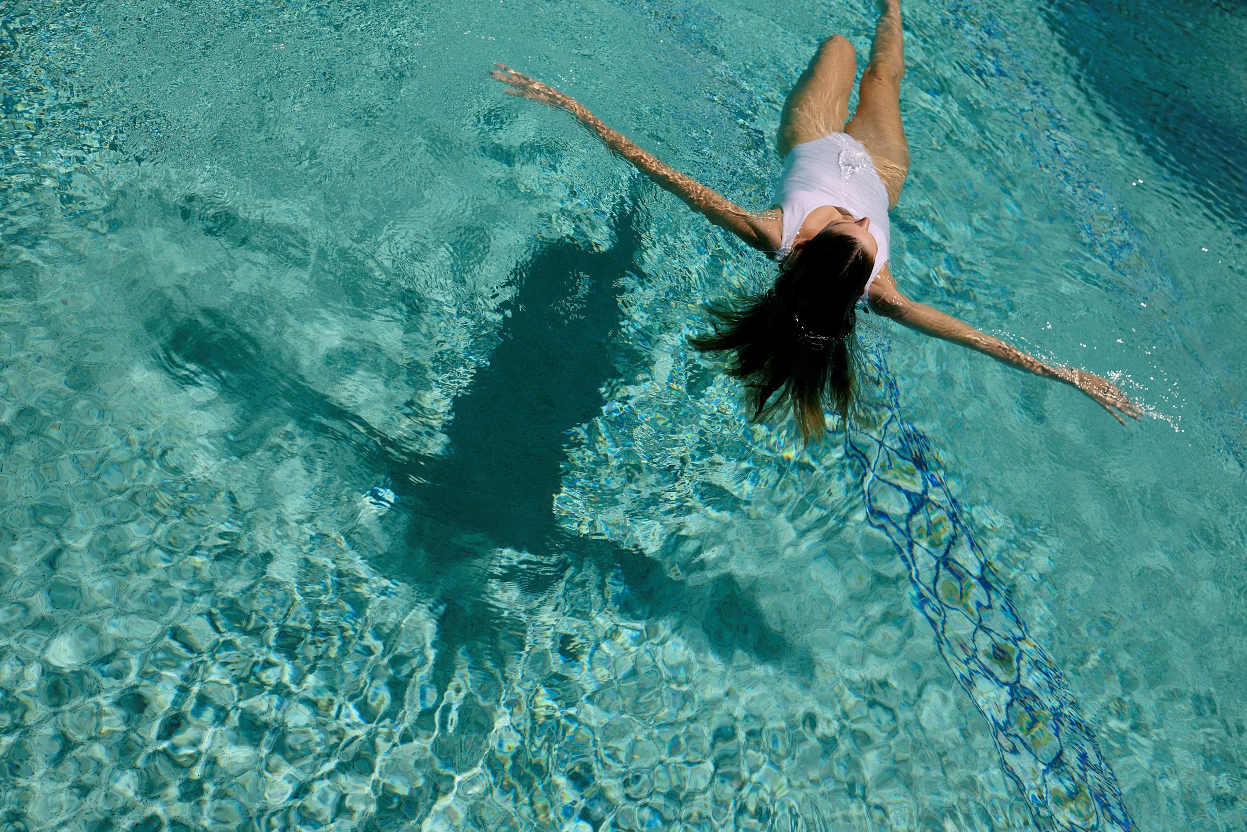 Person floating on their back in a clear blue pool with a large dark shadow beneath the water nearby.