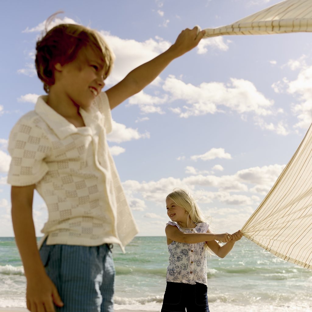 Two children playing with a striped fabric on a beach, with ocean waves and sky in the background.