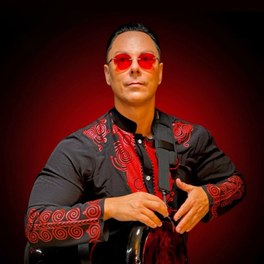 Person in patterned black shirt and red‑tinted sunglasses holding a stringed instrument against a dark red background.
