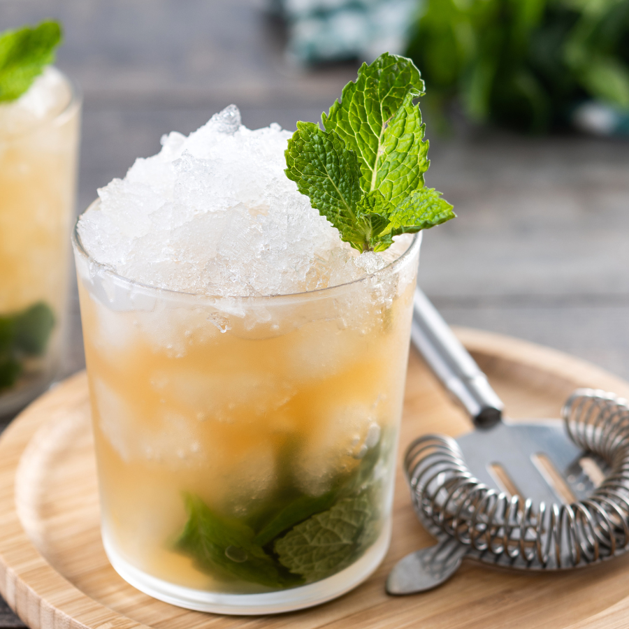 Glass of crushed‑ice cocktail with fresh mint garnish on a wooden tray beside a metal strainer.