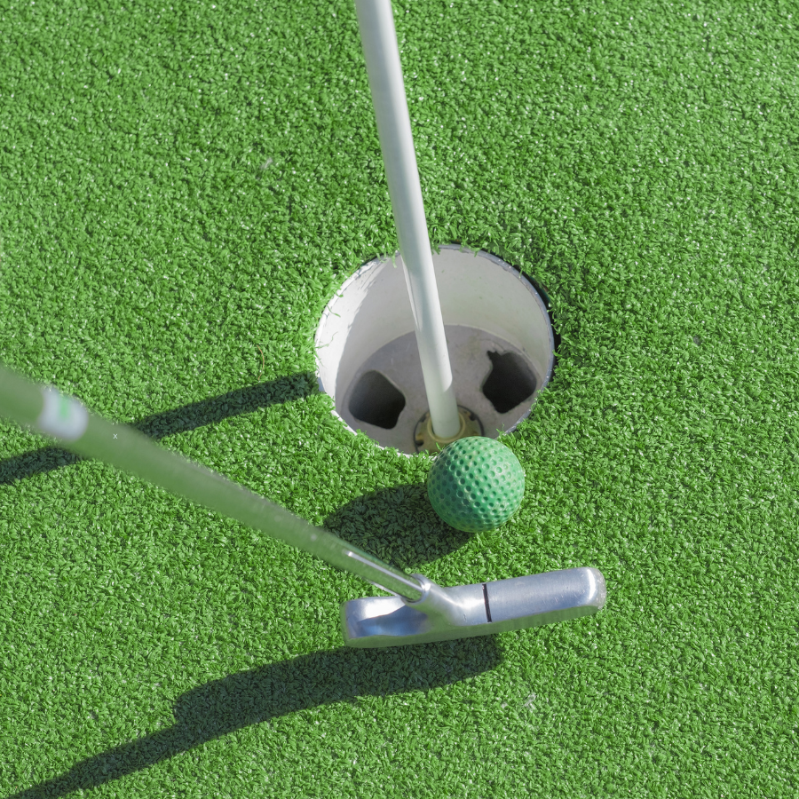Green mini‑golf ball beside a hole with a putter on bright green turf.