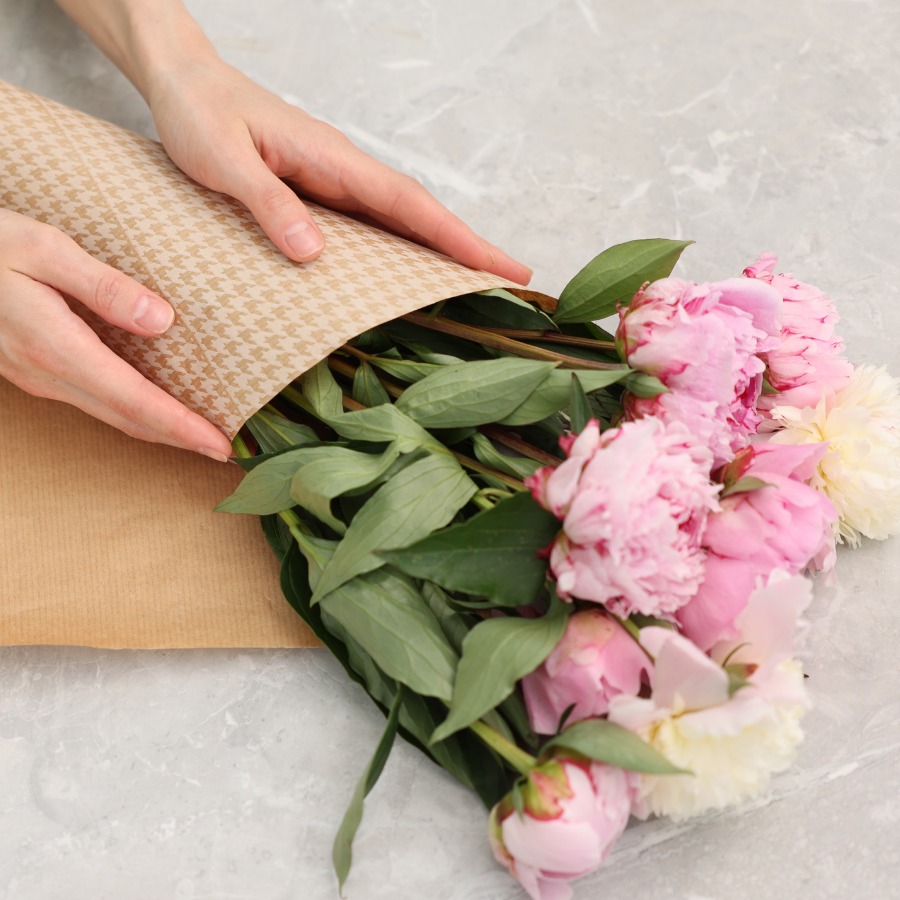 Hands wrapping a bouquet of pink and white peonies in patterned brown paper on a marble surface.