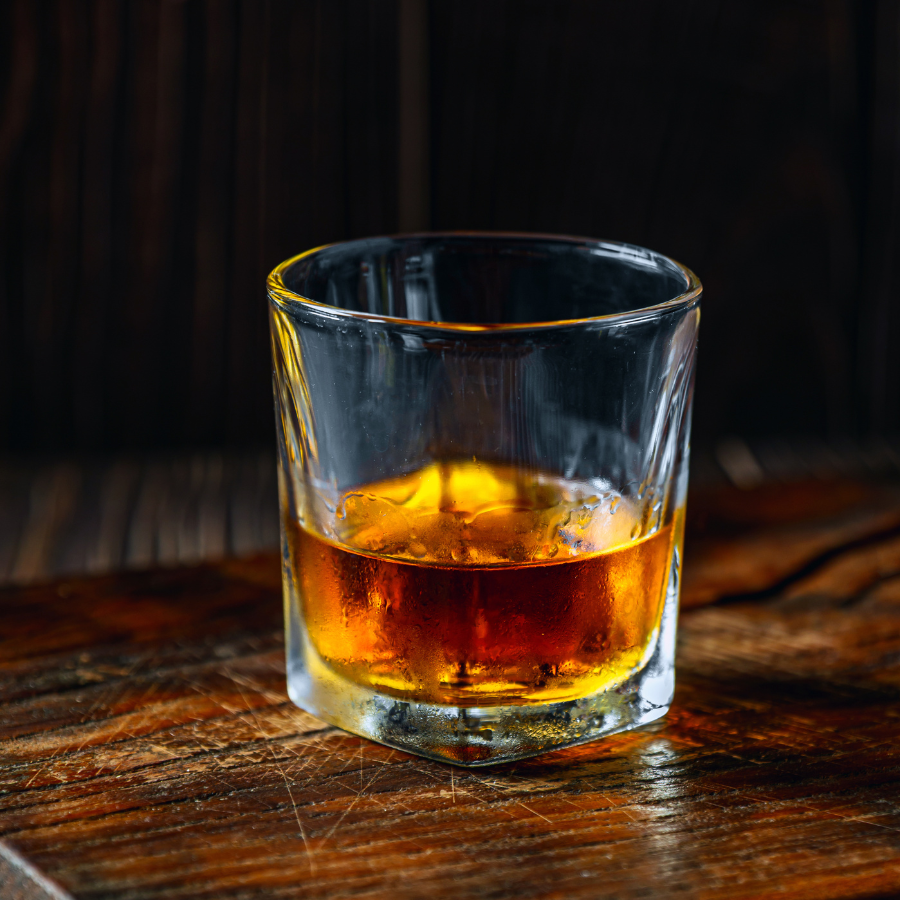 Glass of dark amber liquor with ice on a wooden surface against a dim, rustic background.