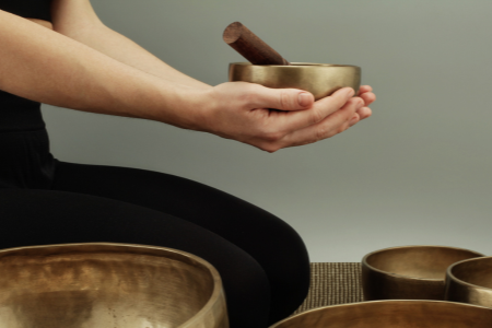 Person seated on the floor holding a brass singing bowl, with bowls arranged on a mat in front against a plain background.